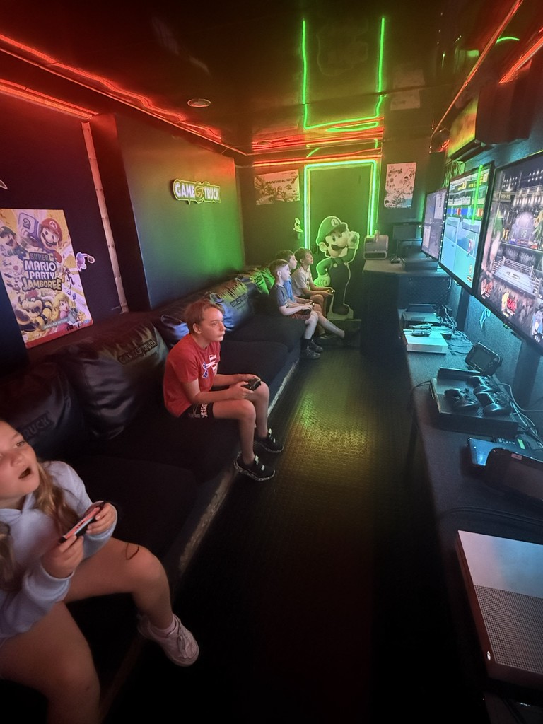 nterior of a mobile gaming theater with vibrant neon green and red LED strip lighting, featuring several children sitting on a black bench playing video games on multiple wall-mounted flat-screen monitors.