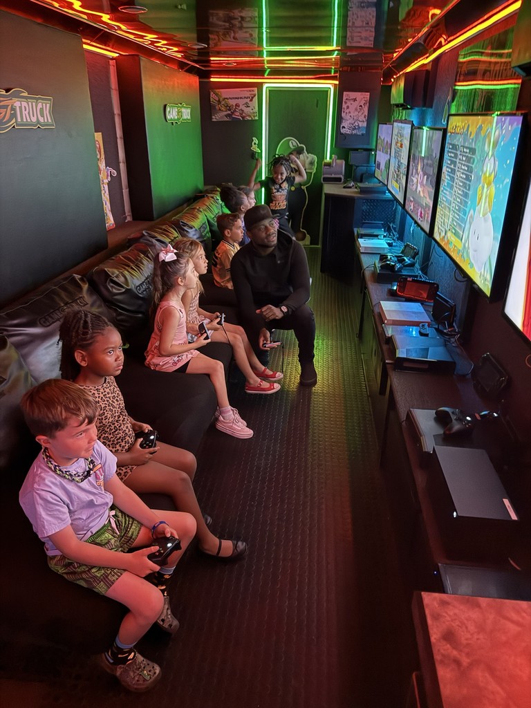 Interior of a mobile gaming theater with vibrant neon green and red LED strip lighting, featuring several children sitting on a black bench playing video games on multiple wall-mounted flat-screen monitors.