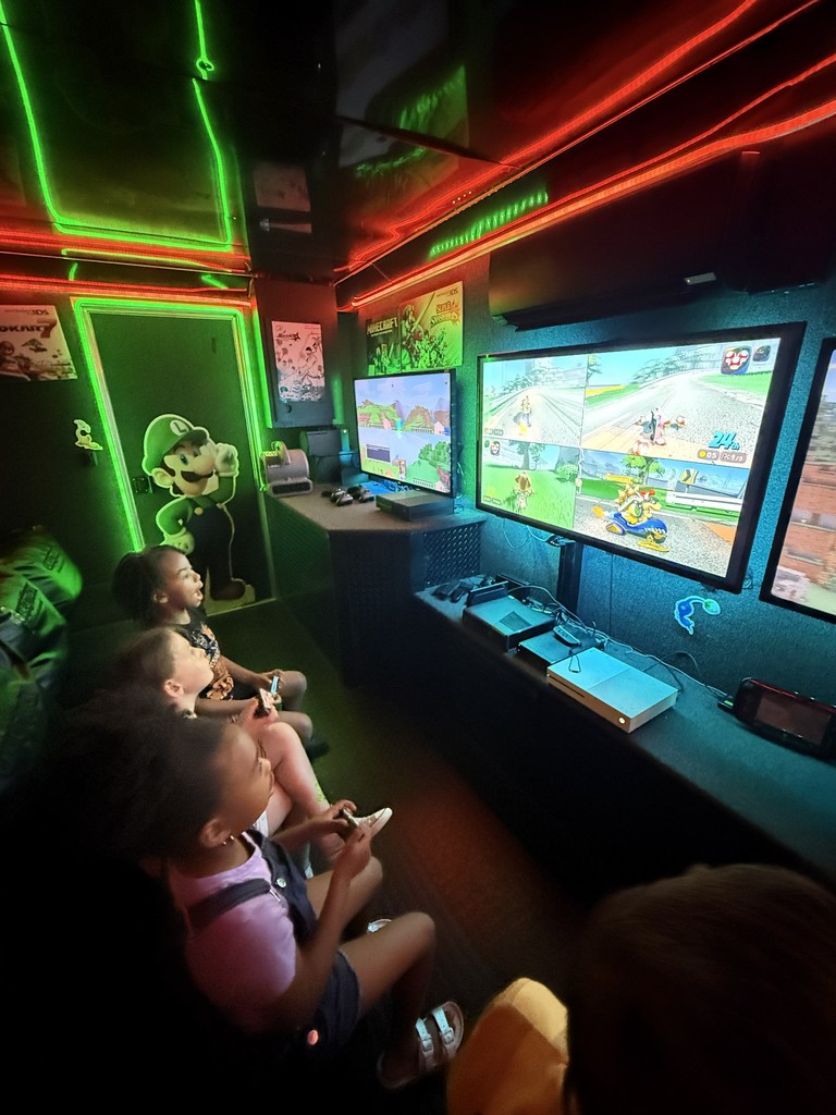 nterior of a mobile gaming theater with vibrant neon green and red LED strip lighting, featuring several children sitting on a black bench playing video games on multiple wall-mounted flat-screen monitors.