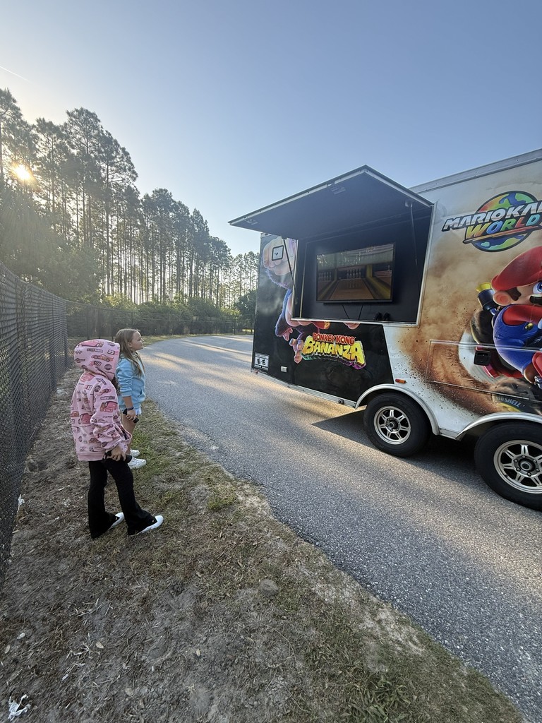 Exterior view of a white "Game On" mobile gaming truck parked on a grassy area near trees, showing the side entrance and colorful graphic wraps.
