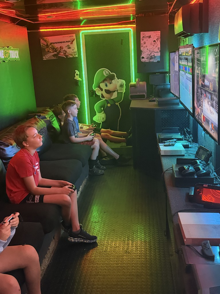 nterior of a mobile gaming theater with vibrant neon green and red LED strip lighting, featuring several children sitting on a black bench playing video games on multiple wall-mounted flat-screen monitors.