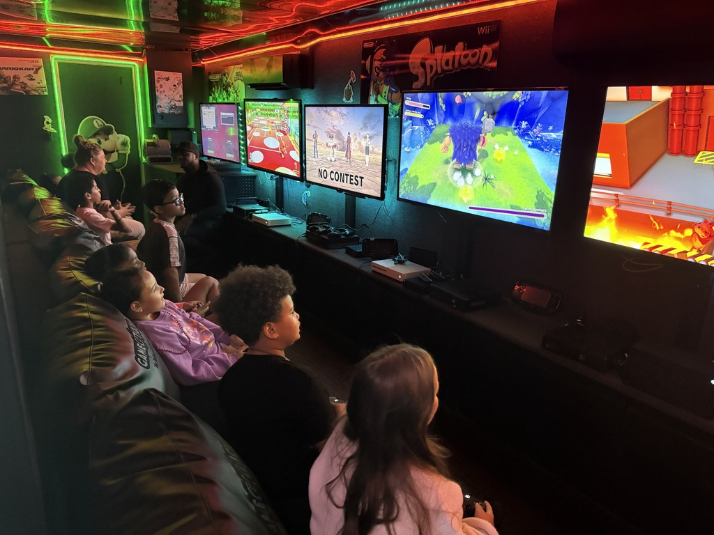 nterior of a mobile gaming theater with vibrant neon green and red LED strip lighting, featuring several children sitting on a black bench playing video games on multiple wall-mounted flat-screen monitors.
