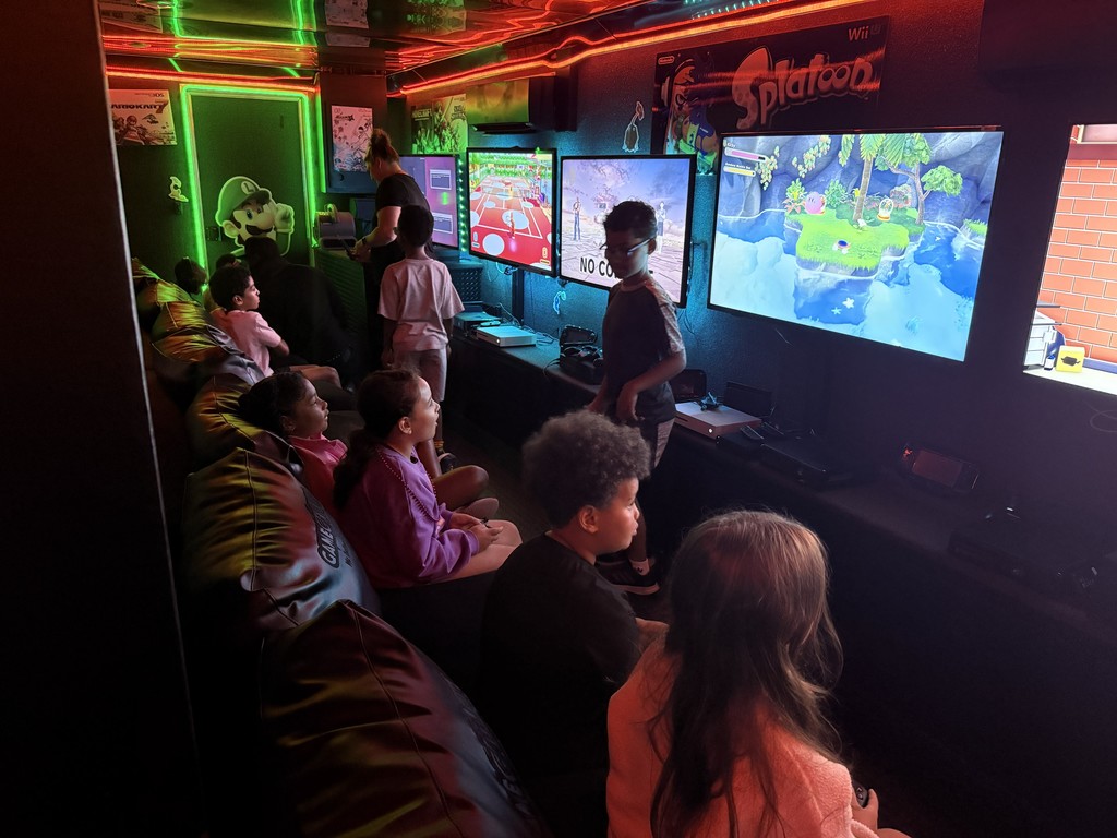nterior of a mobile gaming theater with vibrant neon green and red LED strip lighting, featuring several children sitting on a black bench playing video games on multiple wall-mounted flat-screen monitors.