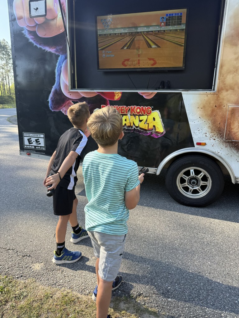 Exterior view of a white "Game On" mobile gaming truck parked on a grassy area near trees, showing the side entrance and colorful graphic wraps.