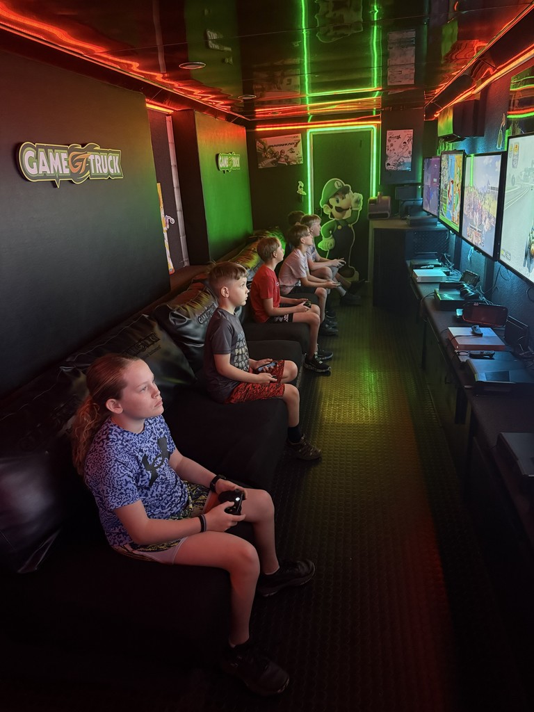 Interior of a mobile gaming theater with vibrant neon green and red LED strip lighting, featuring several children sitting on a black bench playing video games on multiple wall-mounted flat-screen monitors.