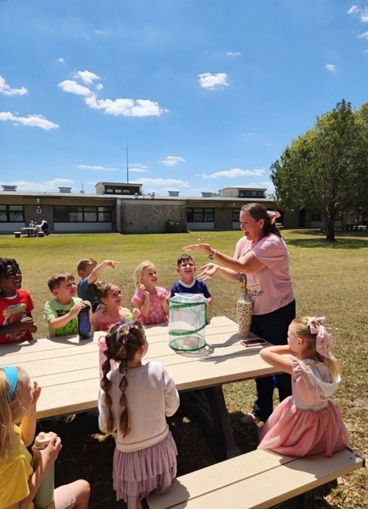 A teacher stands outdoors with a group of young students gathered around a table, demonstrating a butterfly habitat while students watch with excitement.