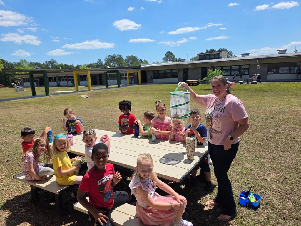 A teacher and a group of elementary students smile outdoors at a picnic table while holding a mesh butterfly habitat used to observe the butterfly life cycle.