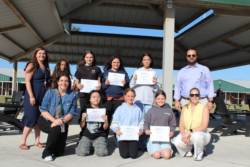 Two art teachers, the principal and assistant principal all pose for the camera under the pavilion with students who won their art competition. Students are holding their certificates and smiling. 