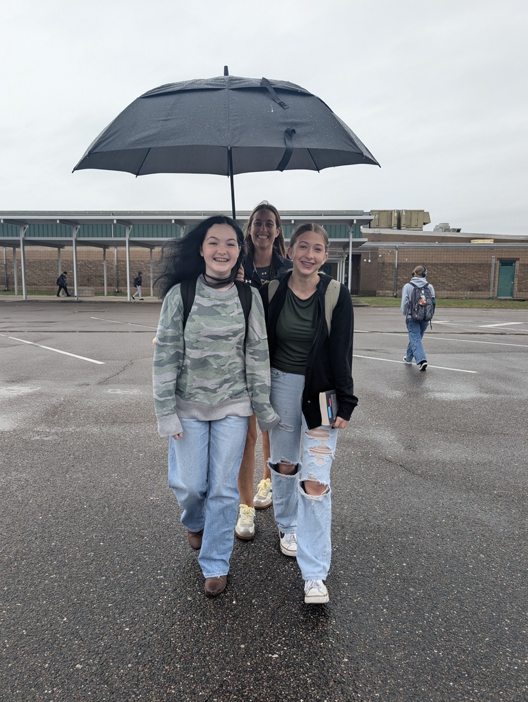 Mrs. Murray walks behind two students as the walk across the bus parking lot to school. She is holding an umbrella over the two students so they don't get wet from the rain. 