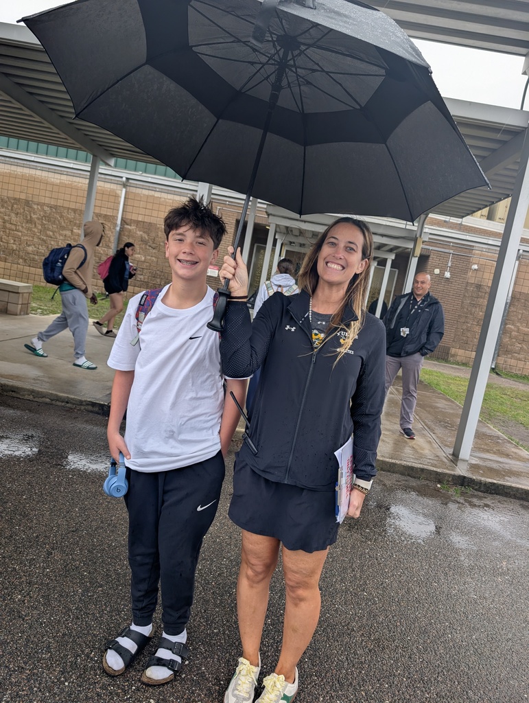 Mrs. Murray stands in the rain with a student, sheltering him from the rain with her umbrella as they wait for a bus to pass by. 