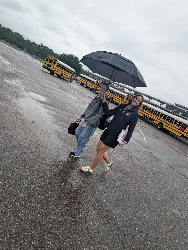 Mrs. Murray and a student walk side by side as she escorts him across the bus parking lot with her umbrella so that he doesn't get wet. 