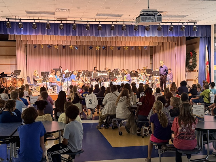 A student band on stage performing in the cafeteria for a group of 5th grade students who are sitting at tables watching the performance. 