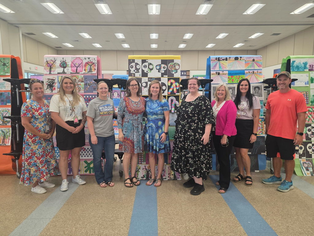A group of nine smiling teachers and volunteers posing together in the school gym, surrounded by vibrant displays of student artwork.