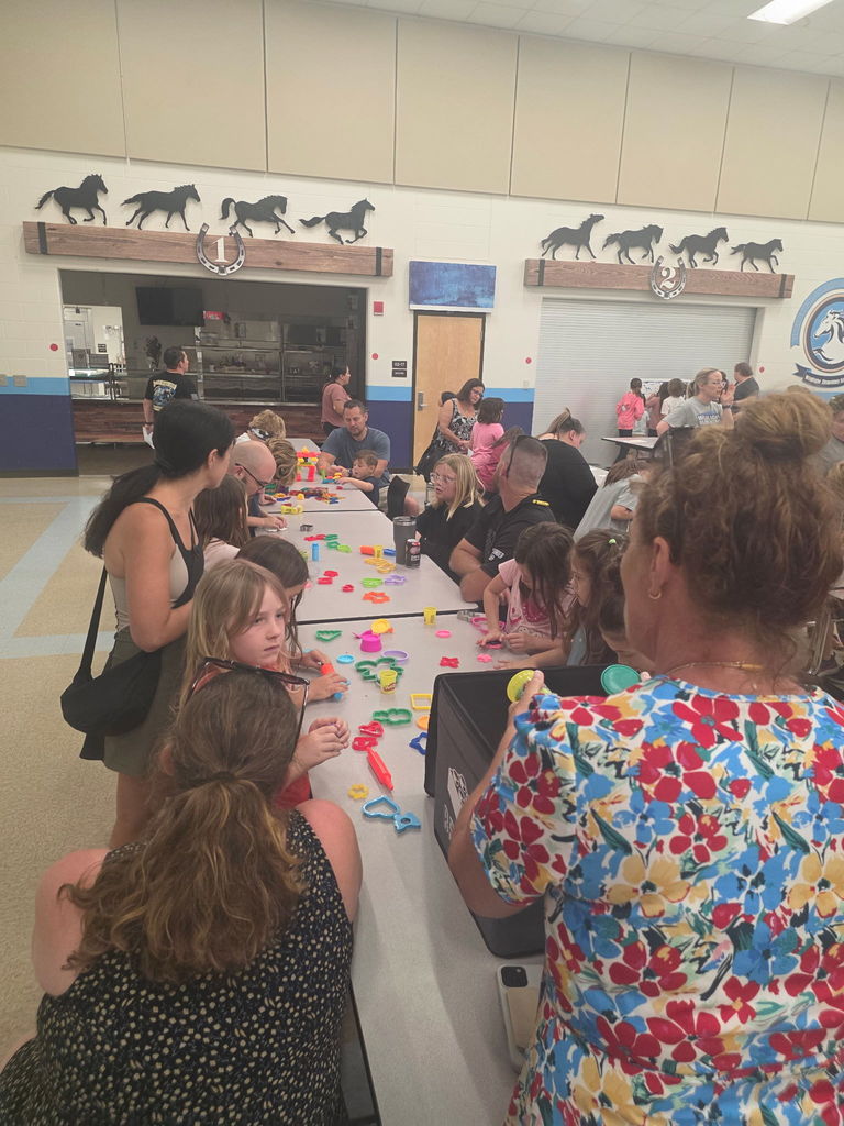 Two volunteers smiling while standing behind a craft table, prepared to assist students with hands-on art activities.