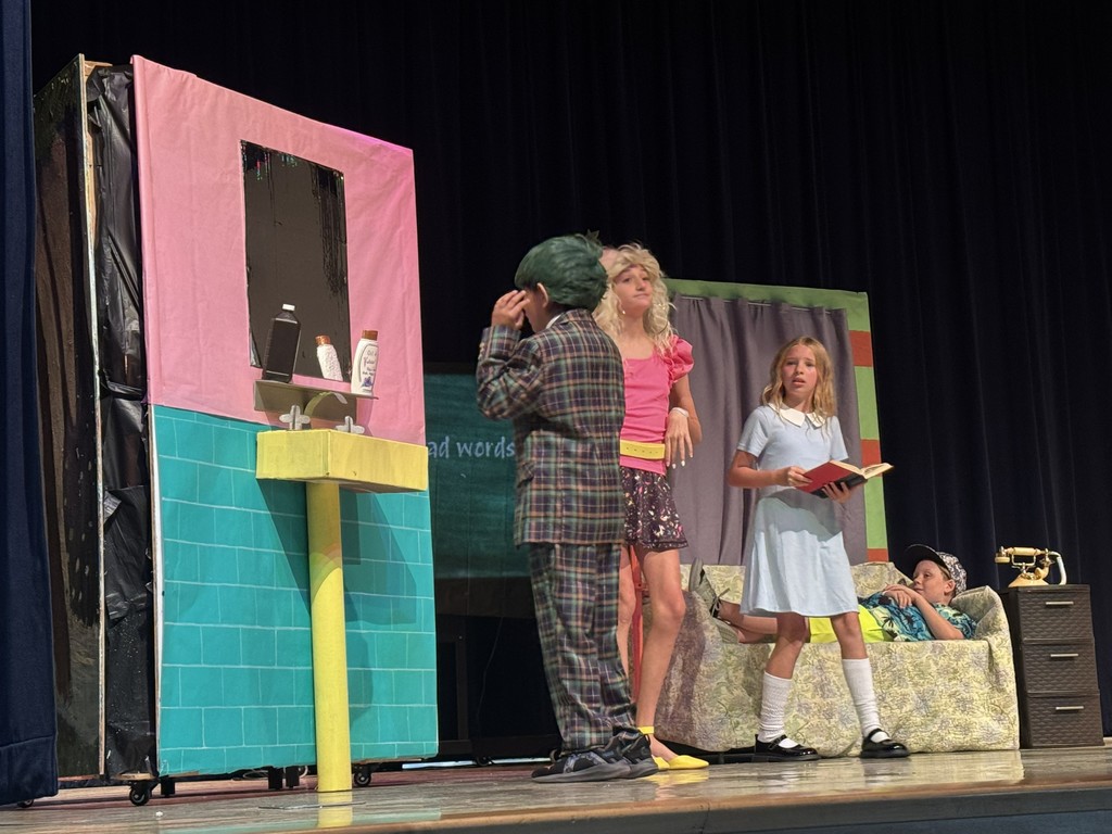 Four students perform a scene from Matilda on stage. One student in a suit and hat stands near a colorful, hand-painted bathroom set piece, while another student in a dress holds a book, and a third student sits on a decorated sofa in the background."