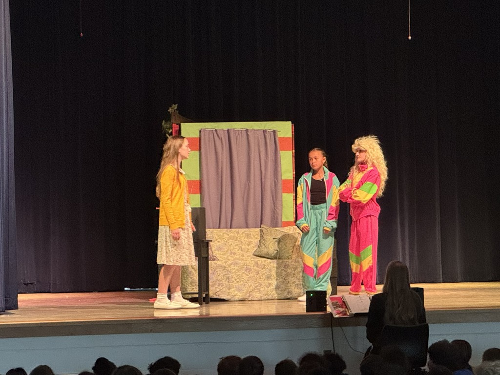 Three students perform on stage in front of a colorful set piece. A student in a yellow cardigan stands to the left, while two others in bright, multi-colored tracksuits stand near a decorated wardrobe.