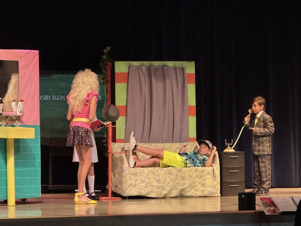 Two students in character costumes interact near the center of the stage. The background features hand-painted set pieces depicting a school or home interior.
