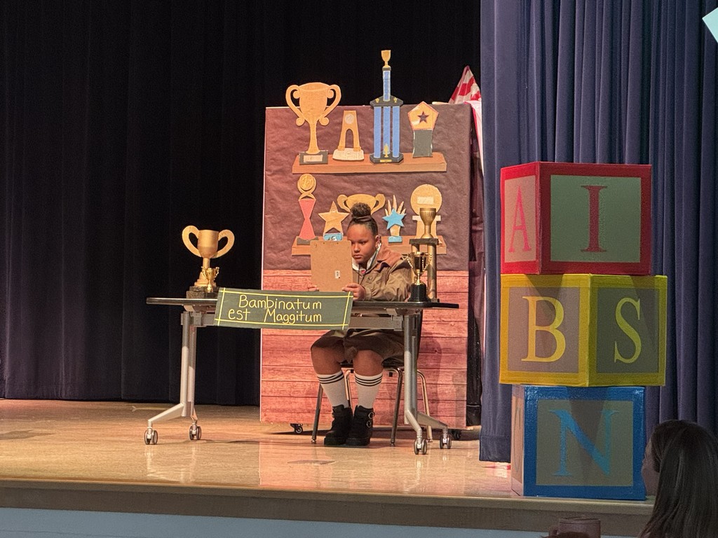 A student performer dressed as Miss Trunchbull sits at a desk on stage during a production of Matilda. The stage is decorated with large colorful alphabet blocks and a backdrop featuring various trophies."