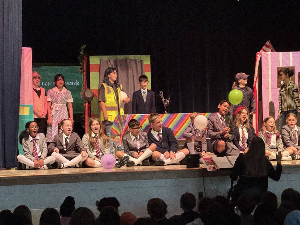 A group of students in school uniform costumes stand in a line across the stage, performing a choreographed musical number under bright stage lights.