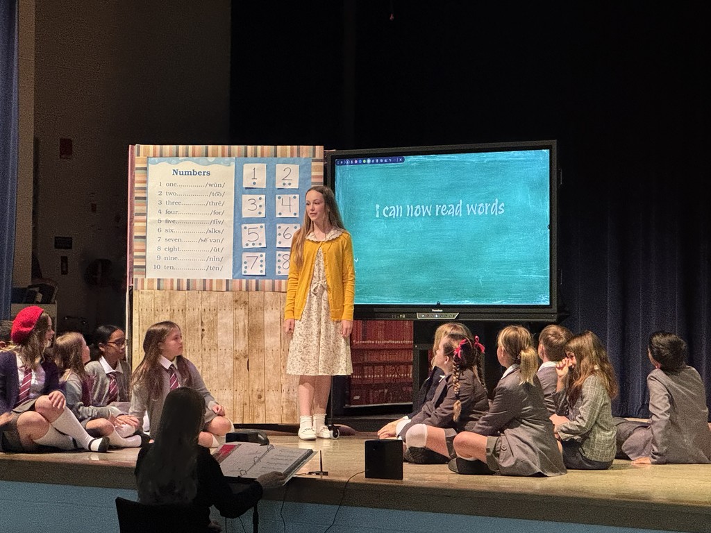 A wide shot of the stage featuring a large group of students. Some are seated on the floor while others stand behind them, creating a layered ensemble scene during the performance.