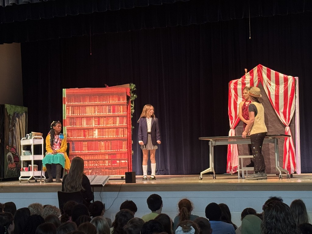 A dynamic scene where several students are gesturing toward a central character, capturing a dramatic moment from the play.