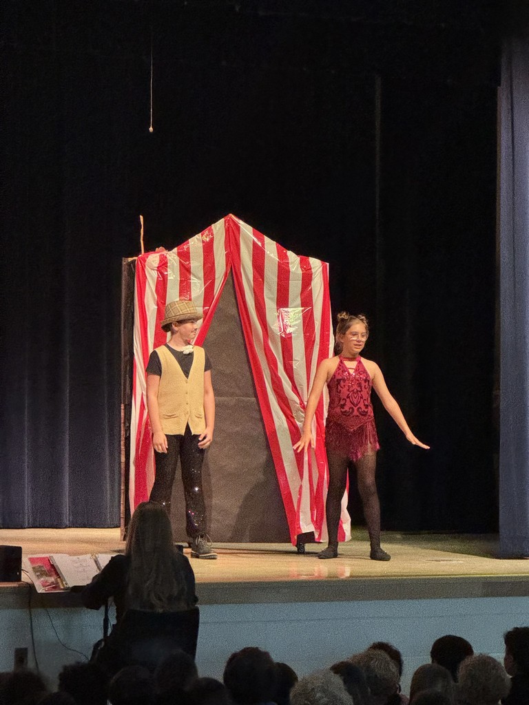 A close-up shot of two lead characters talking on stage, framed by the dark theater curtains and stage lighting.
