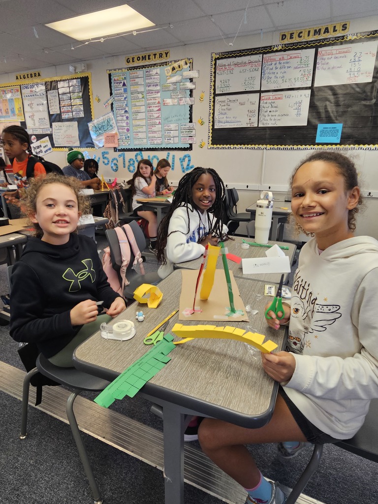 students standing in a classroom, proudly displaying their engineering projects made of yellow and blue building materials.