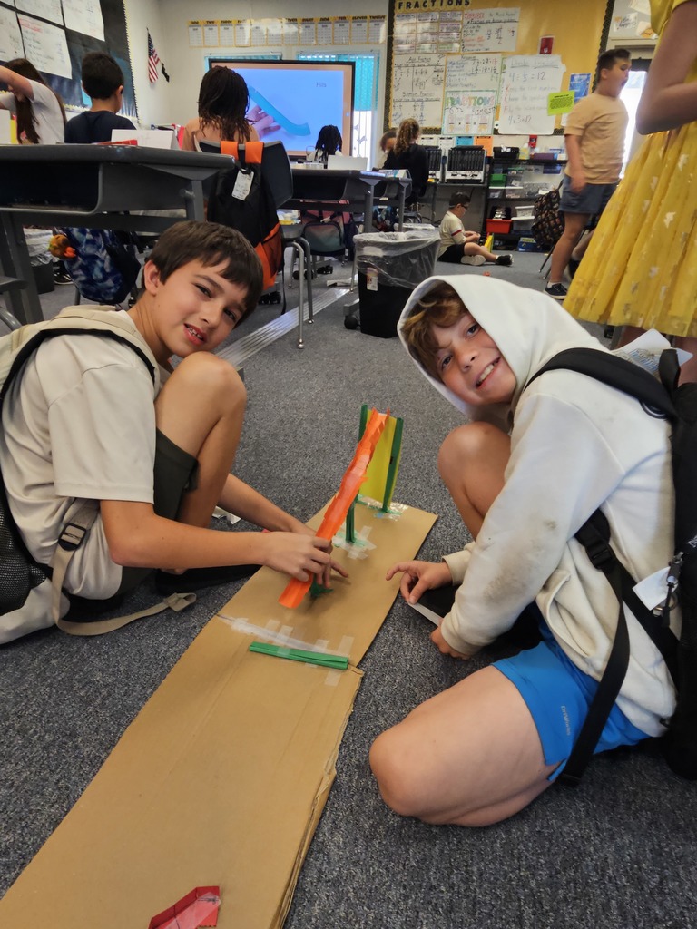 Two students sitting on the floor working together to build a marble run using cardboard, tape, and colorful plastic tracks in a classroom.