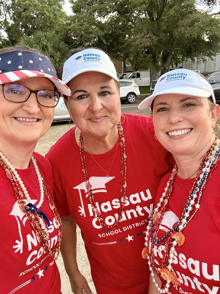 Three Nassau County School District staff members wearing red district shirts and patriotic accessories smile together for a close-up photo outdoors.