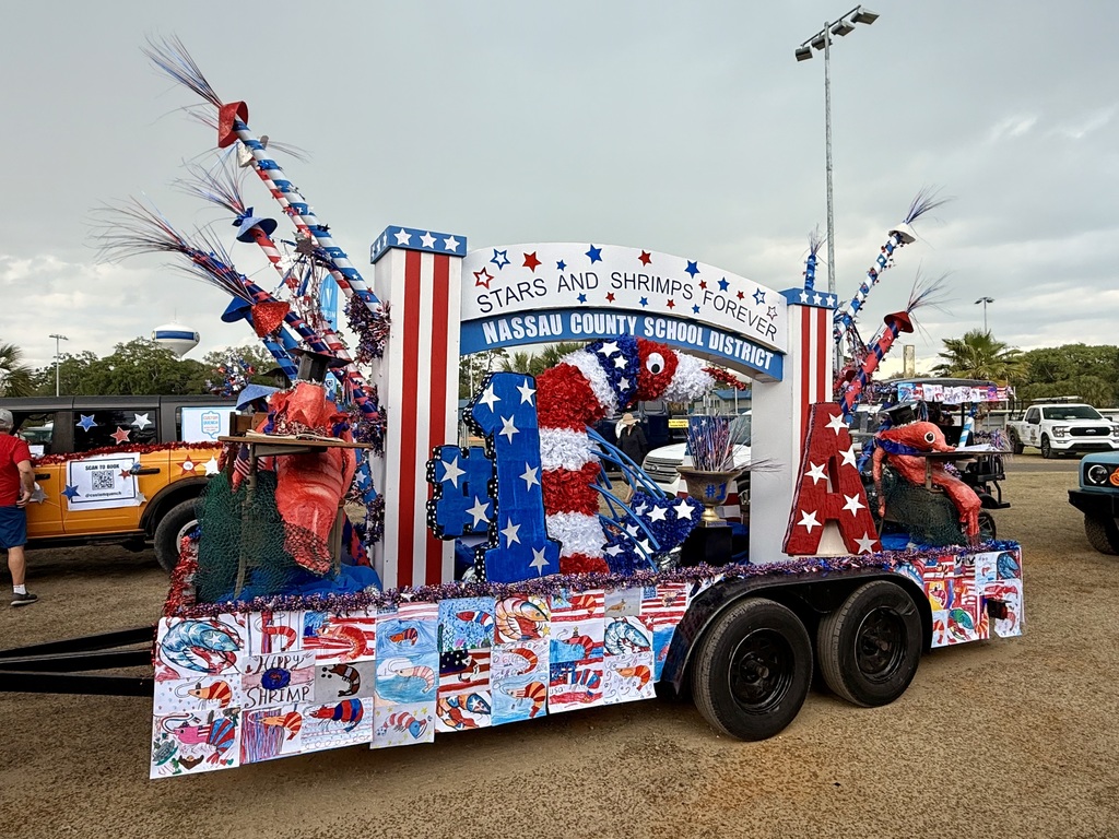 A parade float displays a sign reading “Stars and Shrimps Forever” and “Nassau County School District,” decorated with patriotic colors, a large shrimp figure, and student artwork.
