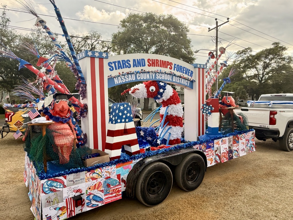 A parade float displays a sign reading “Stars and Shrimps Forever” and “Nassau County School District,” decorated with patriotic colors, a large shrimp figure, and student artwork.