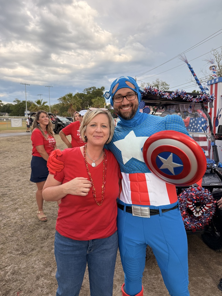 A Nassau County School District staff member stands next to a person dressed as a superhero in front of a decorated parade vehicle.