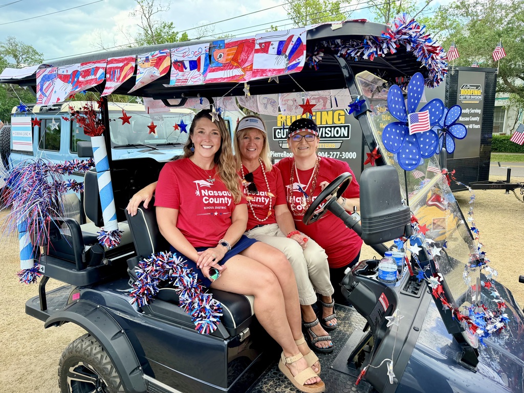 Three Nassau County School District staff members sit on a decorated golf cart with patriotic decorations and smile toward the camera.