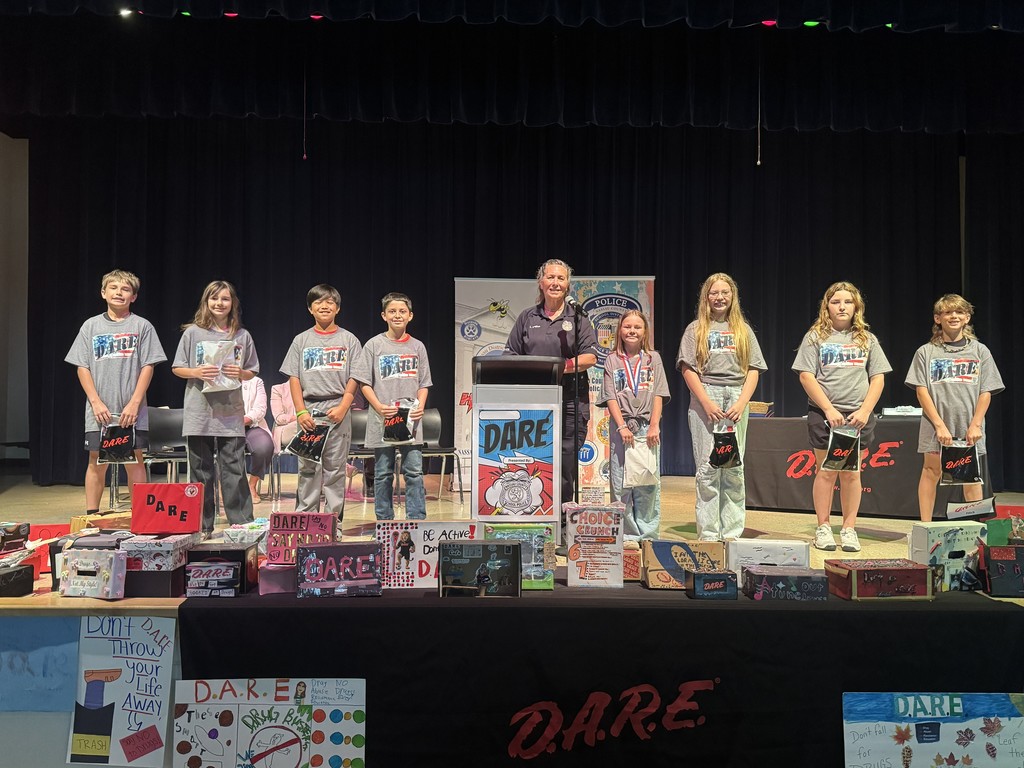 A group of nine 5th-grade students and Officer Laurie Baughhan standing on a stage behind a "D.A.R.E." banner. The students are holding certificates and standing in front of colorful posters they created for the program.