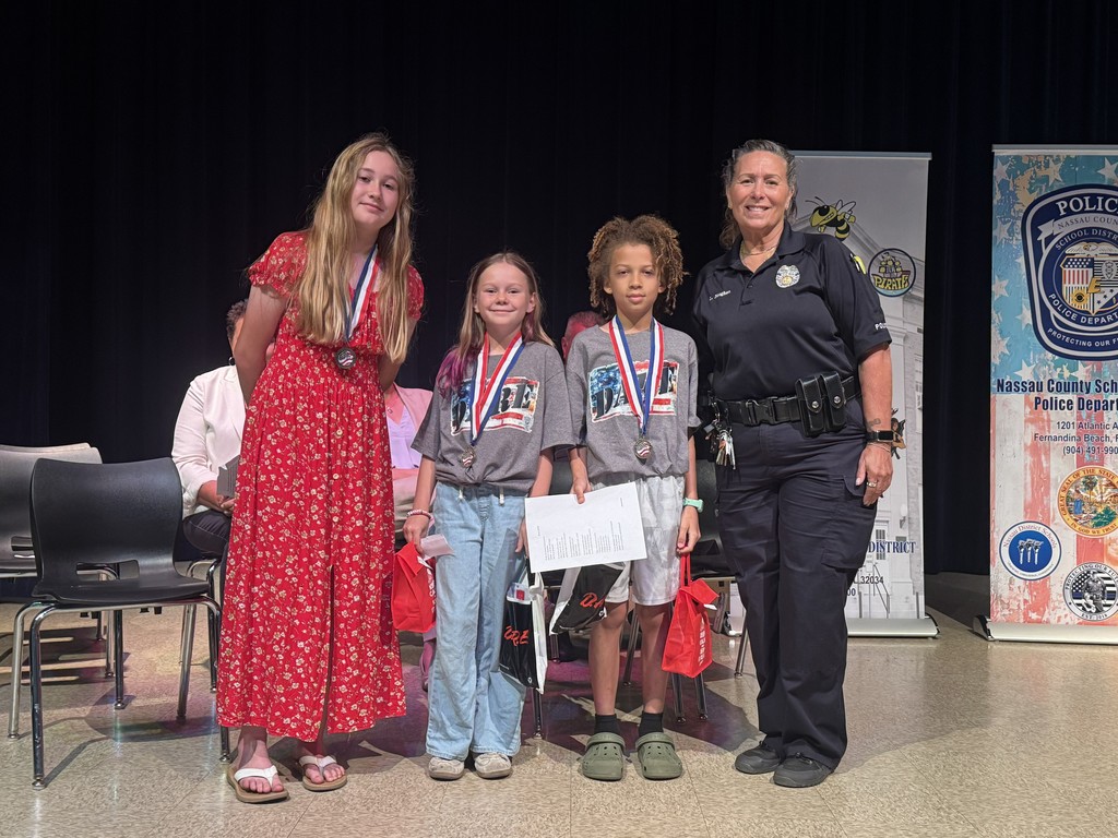 Three students standing on stage with Officer Laurie Baughhan and school administrators. One student is proudly holding up a certificate during the D.A.R.E. graduation ceremony.