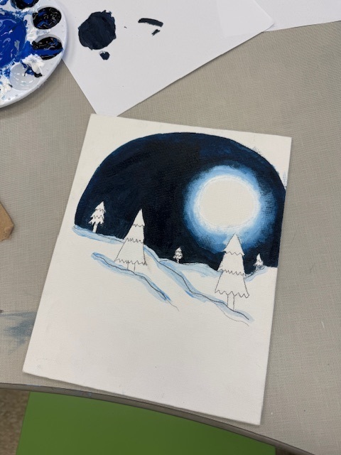 A painted picture lays on a table, half-finished, of a landscape of trees with a black starry night and big full moon on snowy ground. 