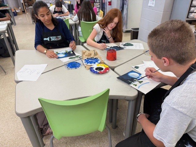 Three students sit at their desks and work on their color scheme paintings.