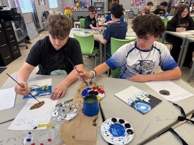 Two students continue to work hard on their color scheme paintings and a table with their brushes and different colored paints. 