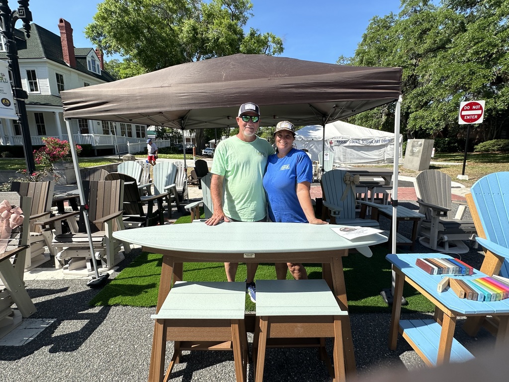 Two adults stand behind a display table under a canopy tent surrounded by outdoor furniture, including chairs and stools, at the Nassau County School District art exhibit area.