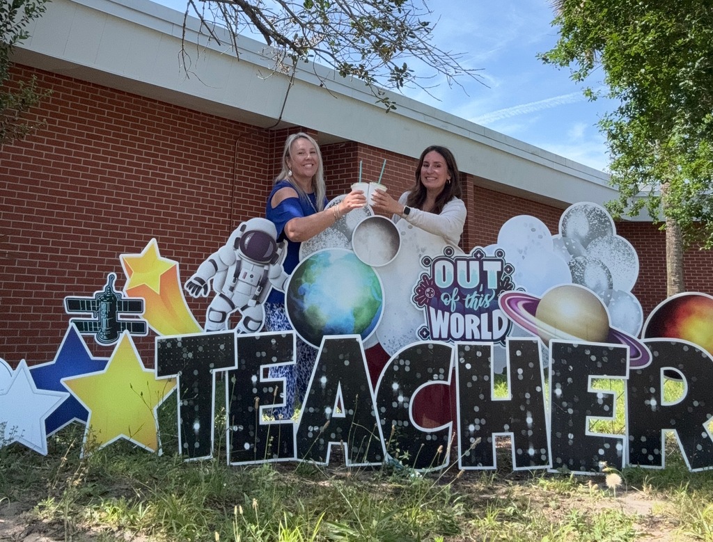 two teachers standing in front of yard card sign saying out of this world holding tropical smoothie smoothies.