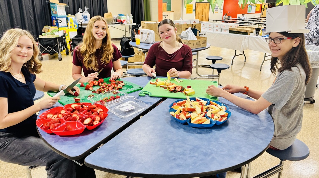 Four students sitting at a table and smile as the cut up fruit for the teacher's morning breakfast.
