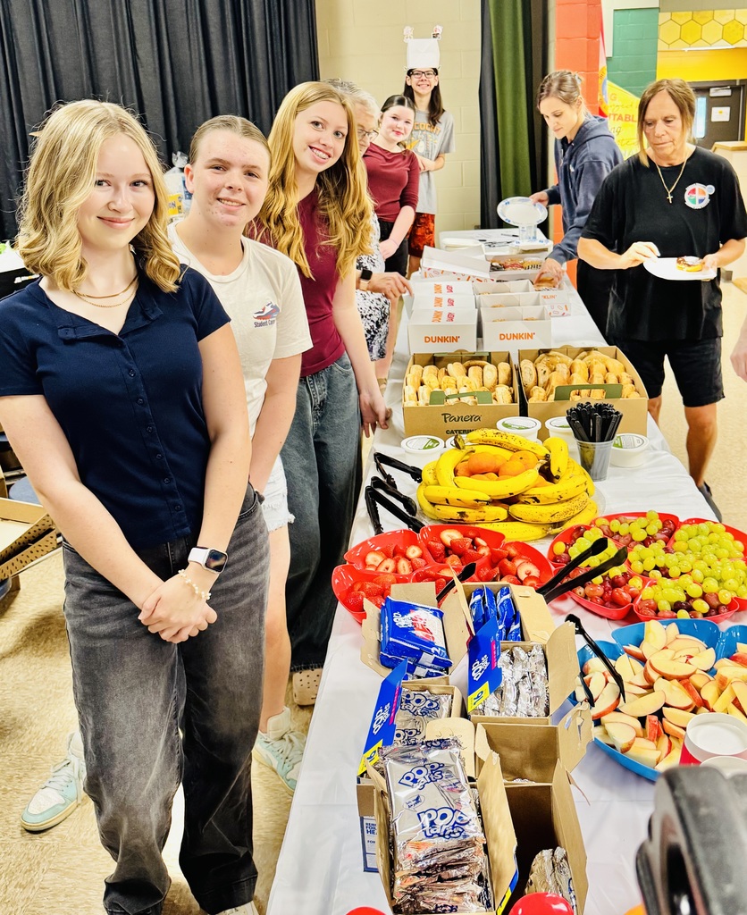 six students lean into the camera photo and smile as they stand next to their long table of breakfast treats that they prepared for teachers and staff. Table is full of pop tarts, fresh fruit, donuts, coffee and bagels. 