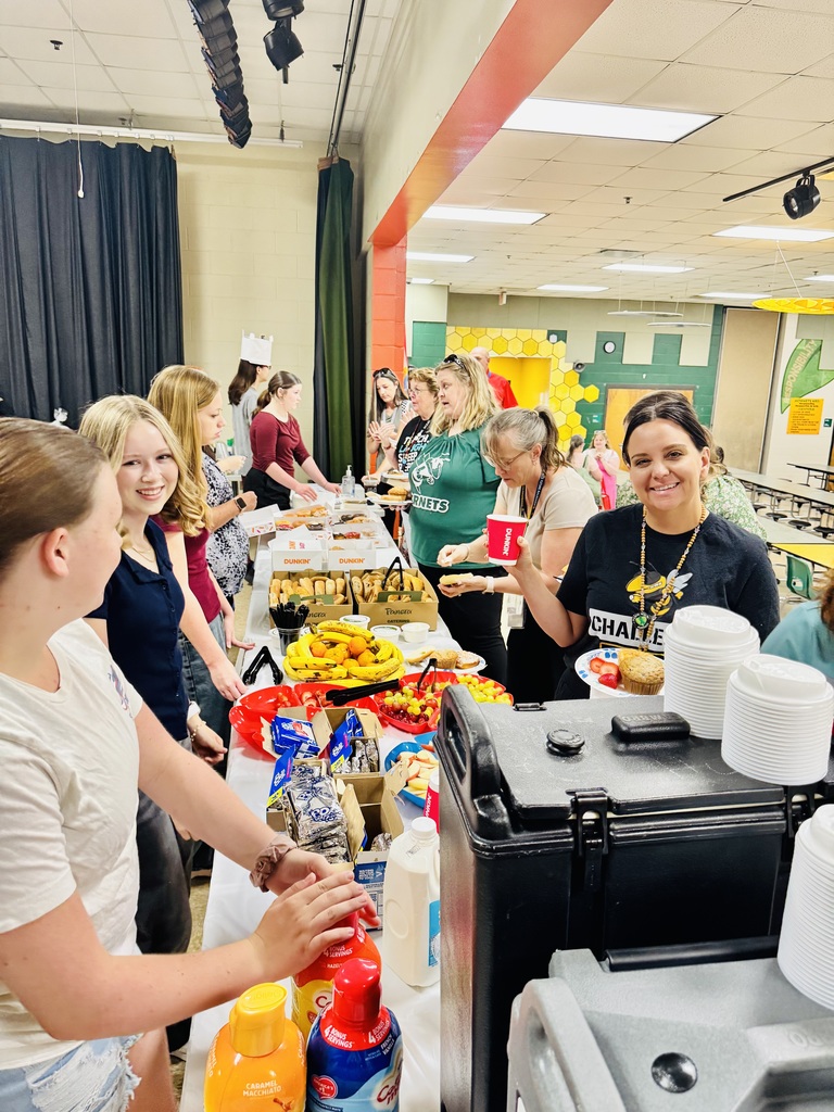 Students serve the teachers a breakfast from a long table with fresh fruit, bagels, coffee and donuts. Teachers lined up on one sid while students serve on the other, all smiles. 