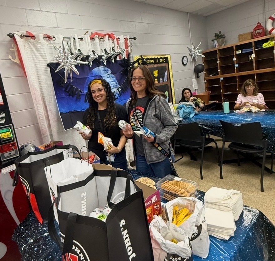 Two teachers standing in front of Jersey Mike's bags full of sandwiches. Both teachers holding sandwiches and bags of chips 
