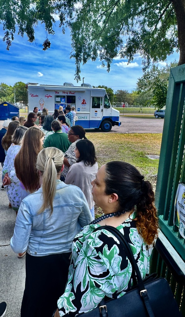 This photo shows a long line of teachers and staff members waiting in line to be served from the Mr. Softee ice cream truck which is shown far away from the foreground of the photo. 
