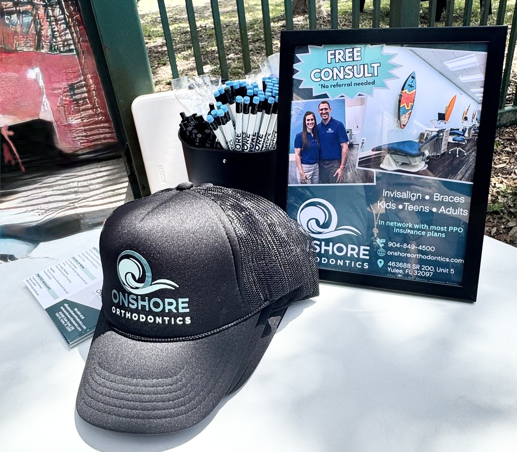 A close up photo of Onshore Merchandise including pens and pencils and handouts and hats. There is also a framed photo with informatoin about a free consult with the Orthodontists, who are married, smiling. 