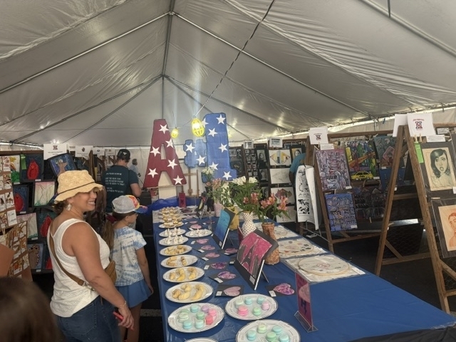 This photo shows the inside of the tent with visitors of the Shrimp Festival browsing the art displays. A blue table holds plates with clay sculptures created by students while on either side drawings and paintings by students are hung up for viewing. 