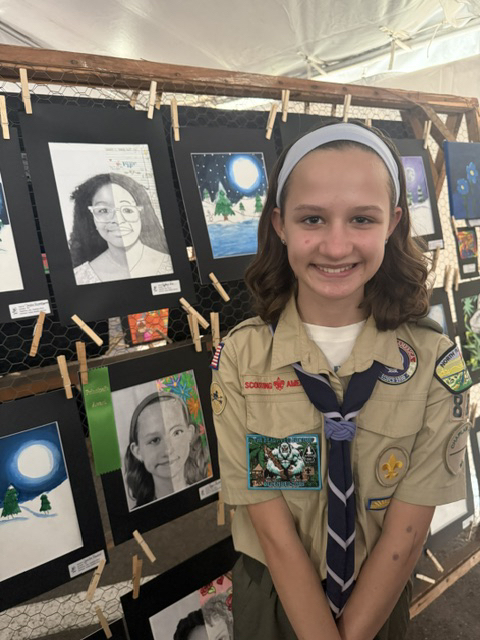A female student smiles for the camera next to her self portrait that is hanging up for viewing in the Nassau County Art Exhibit Tent. 