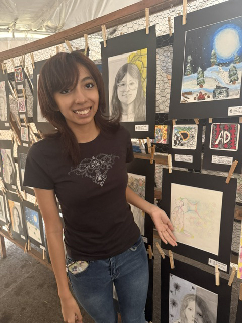 A female student smiles for the camera next to her artistic painting that is being discplayed in the Nassau County Art Exhibit Tent. 