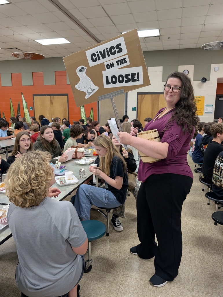 Mrs. Simmons holds a sign that reads, Civics on the Loose! as she walks around the lunch room asking students who are pictured seated at their lunch tables questions about civics and gives out candy to those with correct answers. 